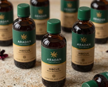 A high-resolution photograph of several bottles of Hibiscus-based products labeled with Aradan Naturals branding. The packaging is sophisticated, using Deep Green and Tan/Gold tones, set against a natural stone surface.