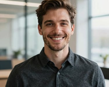 A friendly headshot of a stylish professional man smiling, wearing a modern dark charcoal shirt, set against a bright, airy architectural background in a North American / US creative agency.