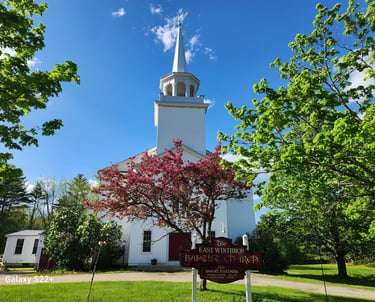 Historic East Winthrop Baptist Church in Maine with a white steeple and blooming pink crabapple tree.