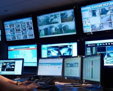 Security professional monitoring multiple live CCTV surveillance camera feeds on a large digital screen wall.