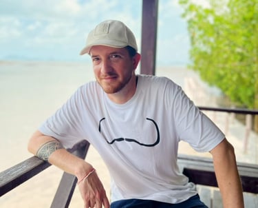 Unicris wearing a white t-shirt and baseball cap sitting on a wooden balcony at a tropical beach.