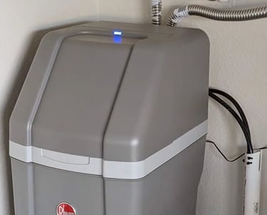 A gray Rheem water softener unit installed against a wall with stainless steel flexible pipes.