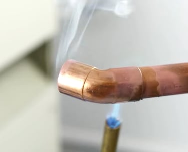 Professional plumber soldering a copper pipe elbow joint with a propane torch and visible smoke.