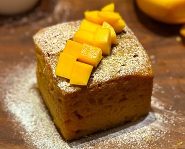 A square slice of mango cake topped with fresh mango chunks and powdered sugar.