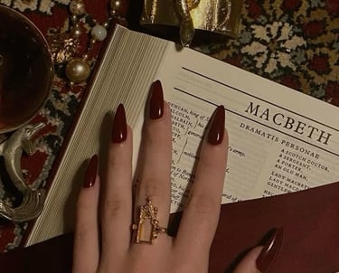 Hand with dark red stiletto nails and gold statement ring resting on Macbeth book.