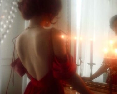 Woman in backless red satin gown facing candlelit mirror, pearl necklace.