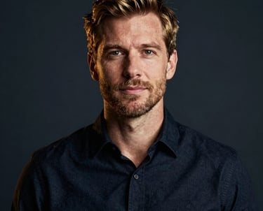 A professional portrait of a man in a modern studio setting. He is wearing a dark navy shirt. The lighting is sophisticated, using burnished gold tones to create depth against a deep charcoal navy background.