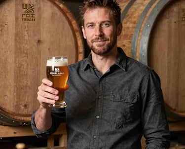 Portrait of our Head Brewmaster in a dark slate shirt, holding a glass of amber-colored Himalayan beer, standing near earthy brown wooden aging barrels.