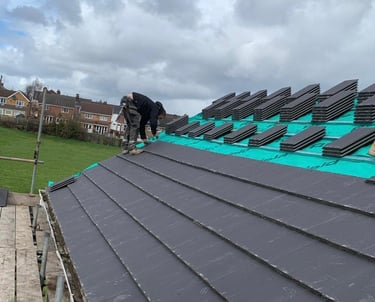 A roofer installs dark grey concrete roof tiles on a house with a green breathable membrane.