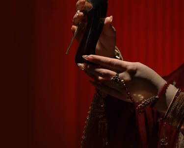 A hand with gold jewelry holding a black high-heel perfume bottle against a red background.