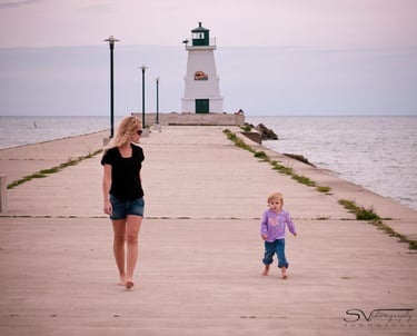 A mother and daughter walking on a pier toward a historic white lighthouse by the sea.