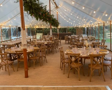 Elegant outdoor wedding reception tent with round wooden tables, bistro lighting, and floral greenery.