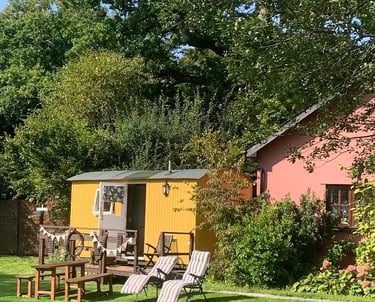 Yellow shepherd's hut and rustic wooden furniture on a sunny lawn beside a pink house.