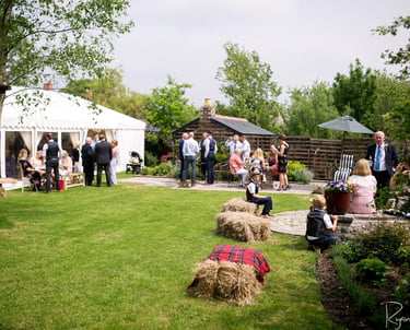Guests mingling at an outdoor garden wedding reception with a white marquee tent and hay bale seating.
