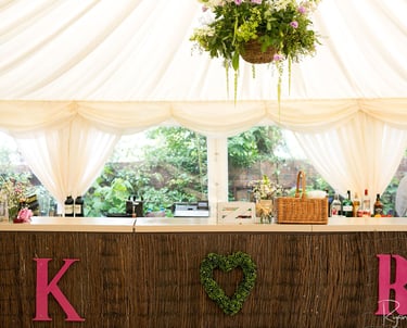Rustic wedding bar setup with pink initials and a heart wreath under a white marquee tent.