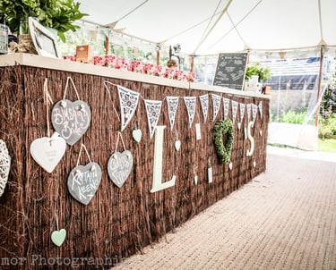 Rustic outdoor wedding bar with heart-shaped signs and white bunting under a marquee tent.