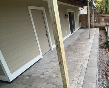 Custom stone paver patio installation under a wooden deck with tan siding and sliding glass doors.