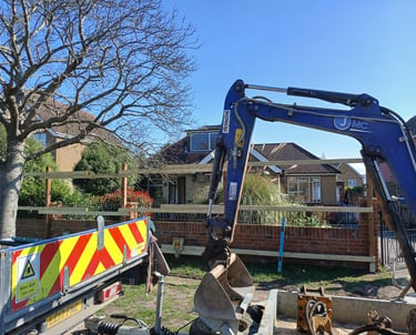 Blue Hitachi mini excavator and flatbed truck at a residential construction site near a wooden fence.
