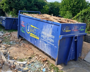 A blue JMC roll-on roll-off skip filled with wood waste on a demolition site with rubble.