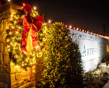 A lit Christmas wreath with a red bow hangs on a stone wall next to sign.