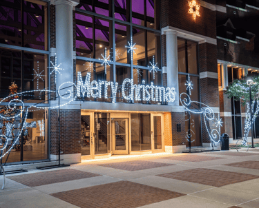 Merry Christmas neon sign and white holiday lights decorating a brick building entrance at night.