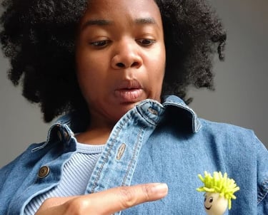 A woman in a denim jacket points at a small smiley face toy in her pocket with a surprised expression.