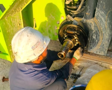 A mechanic in a white hard hat performs heavy equipment brake repair on a construction vehicle axle.
