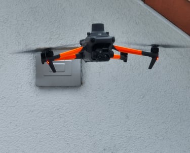A DJI Mavic 3 Enterprise drone with orange skins hovering near a white building wall.