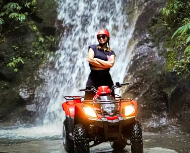 A smiling woman in a red helmet posing on a red ATV quad bike in front of a tropical jungle waterfall.