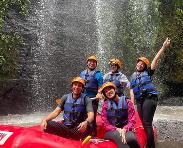 A group of friends in helmets and life jackets posing in a red white-water raft near a waterfall.