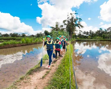 A group of tourists wearing life jackets and helmets walking along a path in Bali rice fields.