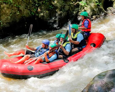 A group of people wearing helmets white water rafting in a red boat on a tropical river.