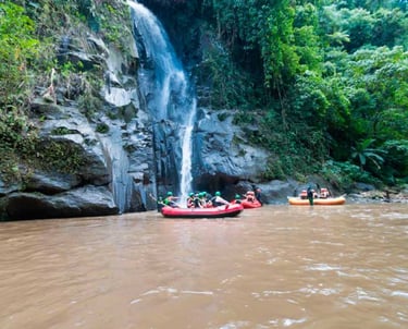 A group of people white water rafting in red boats past a tropical jungle waterfall.
