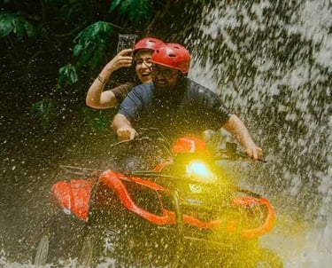 Two people wearing helmets ride a red ATV through a tropical waterfall during an outdoor adventure tour.