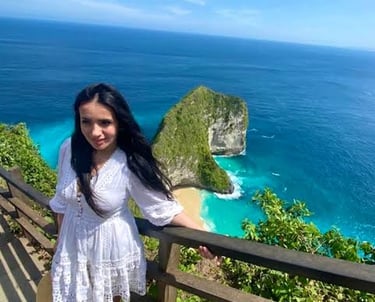 A young woman in a white lace dress poses at Kelingking Beach viewpoint in Nusa Penida, Bali.