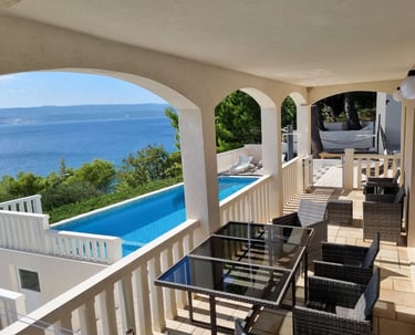 The photo shows an apartment balcony with a dining set and a view of the pool and the sea.