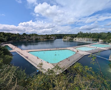 Outdoor swimming pools with turquoise water built on a lake, surrounded by trees and rocky cliffs.