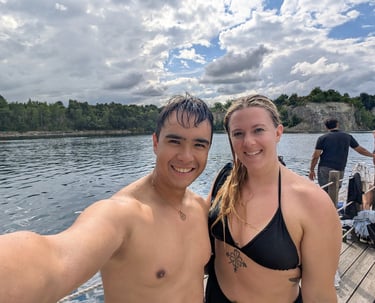 Smiling couple in swimwear taking a selfie by a lake with trees and cloudy sky in the background.