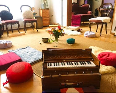 ein Raum mit Stuhlkreis und Sitzkissen, einem Harmonium und Blumenstrauß in der Mitte