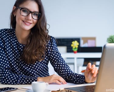 a woman in glasses and a polka dot shirt is smiling and holding a pen with a laptop on the desk