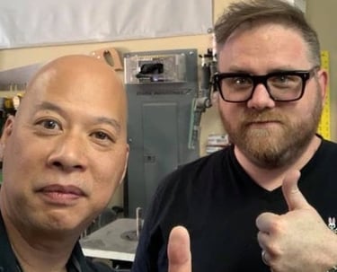 Ray Lum and Josep giving a thumbs up in a workshop setting with woodworking tools in the background.