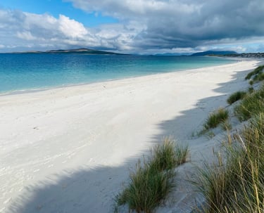 White sand beach with turquoise ocean water and grassy dunes under a cloudy sky in the Outer Hebrides.