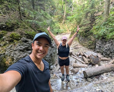 Two hikers smiling and posing on a rocky trail in a forest with fallen logs and greenery around