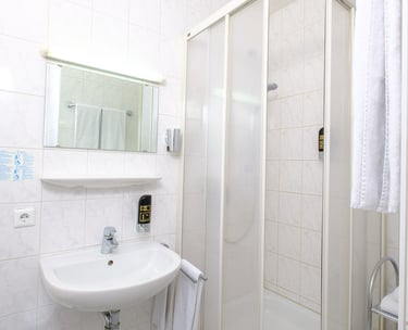 Compact hotel bathroom featuring a white pedestal sink, mirror, and a glass shower stall with white tile walls.