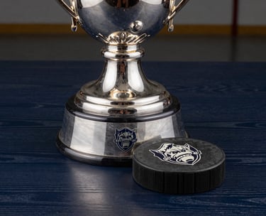 Close-up of a silver collegiate hockey trophy on a dark navy wooden table, etched logos catching the studio light, professional and prestigious feel.