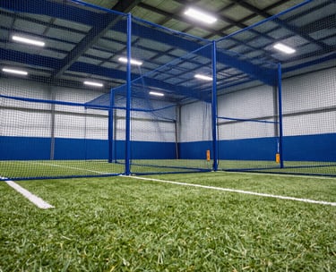 A low-angle shot of a high-performance softball training facility in the North American / US Midwest, showing indoor hitting tunnels with blue netting and professional turf, under bright, clean white lighting.