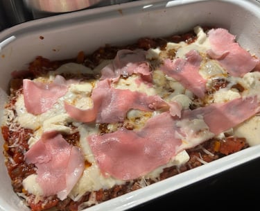 Homemade lasagna in a white baking dish topped with tomato sauce, melted mozzarella, and thin ham slices.