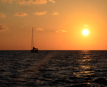 Atardecer, con el cielo anaranjao y un velero de fondo, con las velas recogidas, en medio del mar