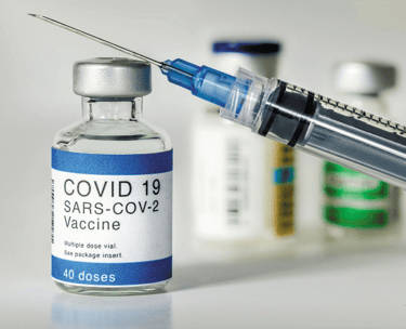 A medical syringe leaning against a COVID-19 SARS-CoV-2 vaccine vial on a white surface.