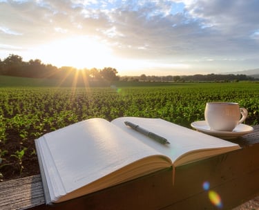 An open journal and a cup of coffee rest on a wooden fence overlooking a farm at sunrise.
